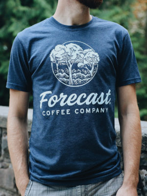 Person wearing a heathered blue Forecast t-shirt in front of a stone wall and trees